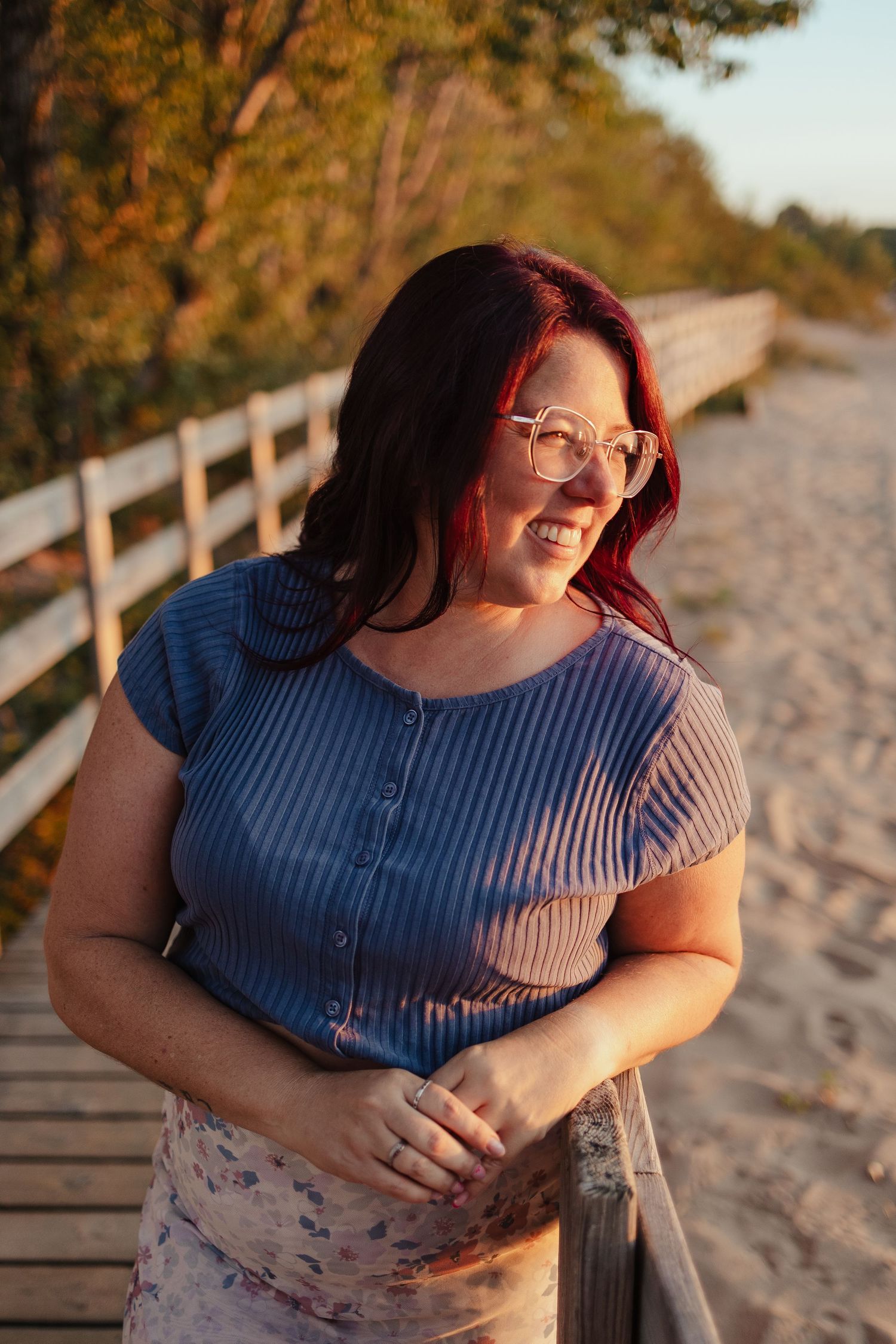 Smiling woman with red hair and glasses leaning on a wooden railing on a sandy boardwalk at golden hour.