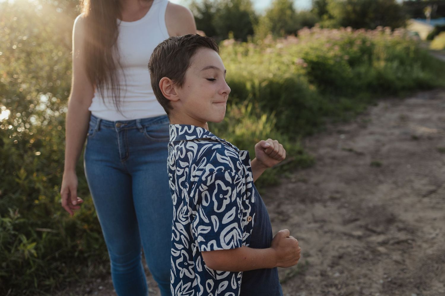 A young boy in a patterned shirt smiles outdoors near a woman in jeans and white top during golden hour.