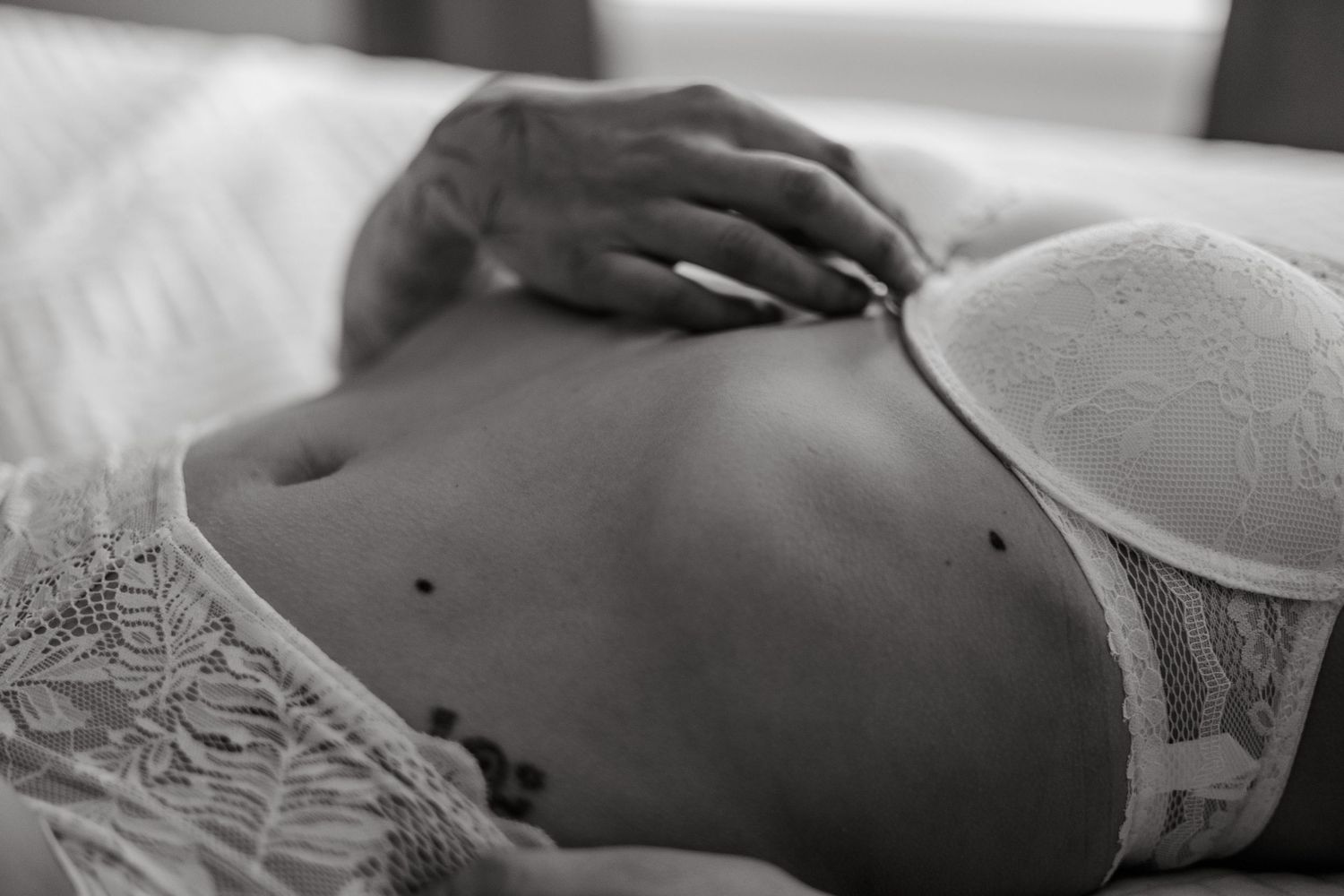 Black and white boudoir photo of a woman in white lace lingerie lying on a bed.