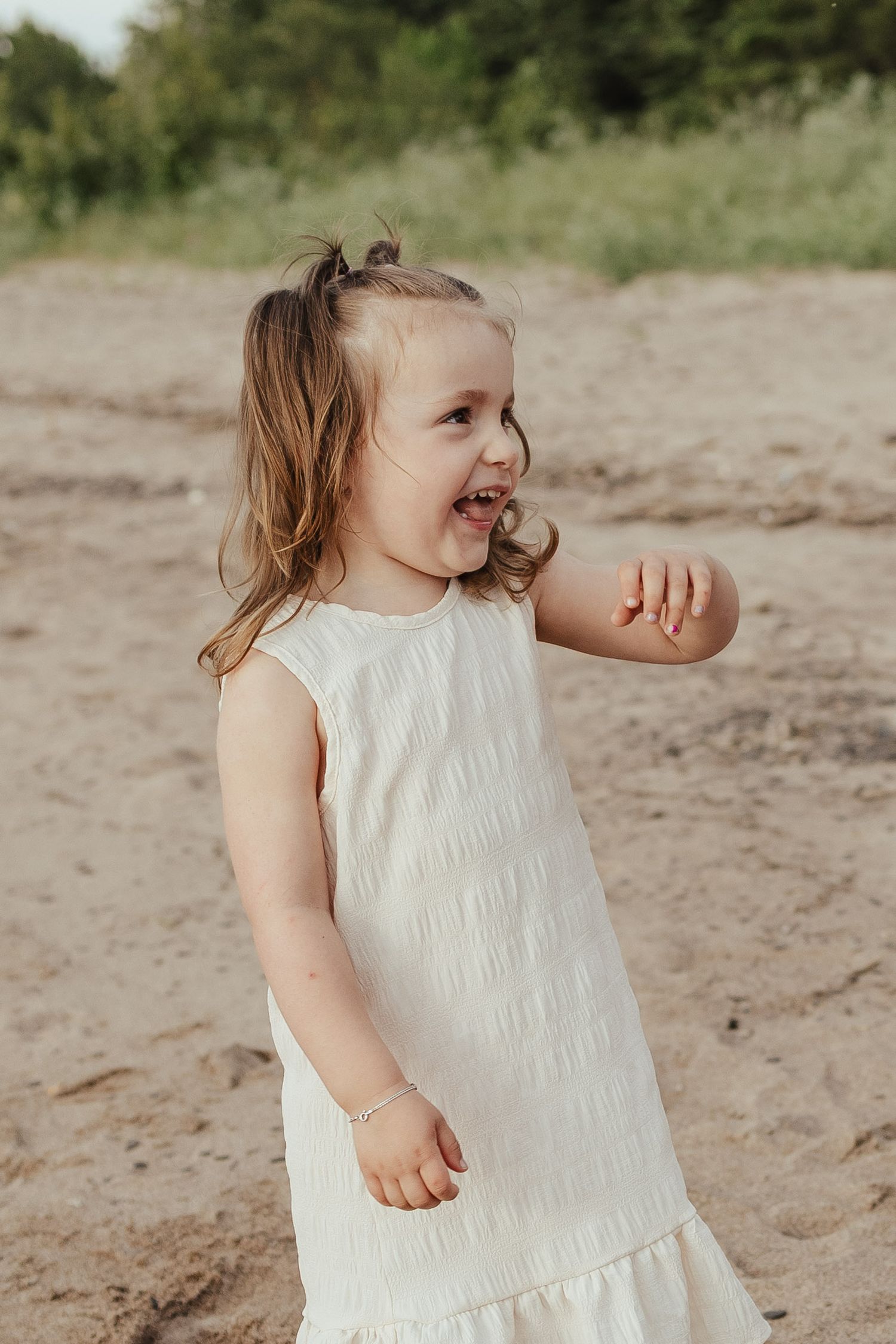 A joyful toddler girl in a white dress laughs and plays on a sandy beach with green trees behind her.