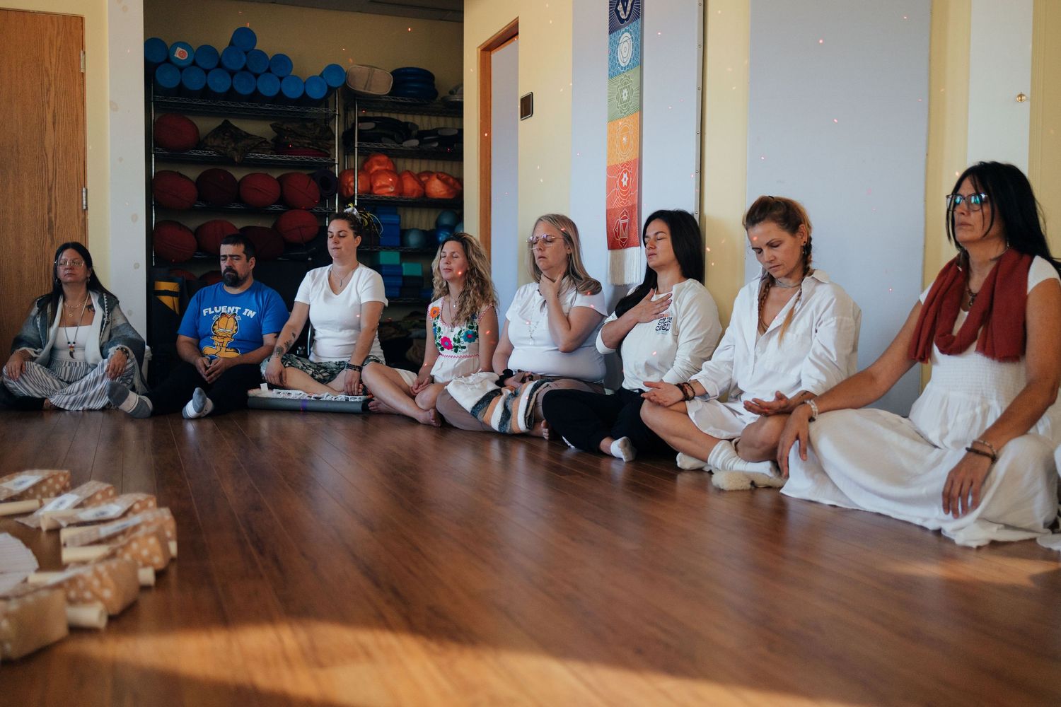 Group of people sitting cross-legged on floor in white clothing during a yoga or meditation session in a studio.
