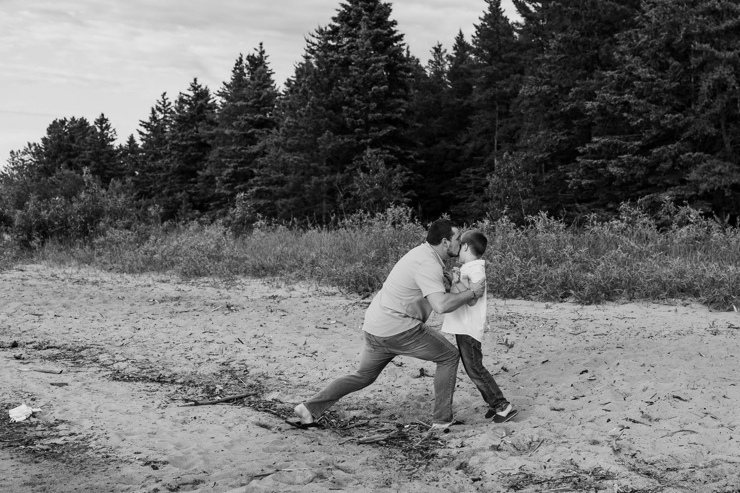 Black and white photo of a parent and child playing together on a sandy beach with evergreen trees in the background.