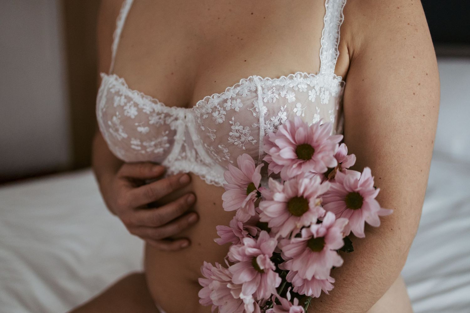 Woman in white lace bra holding pink daisies bouquet, elegant boudoir photography with soft natural lighting.
