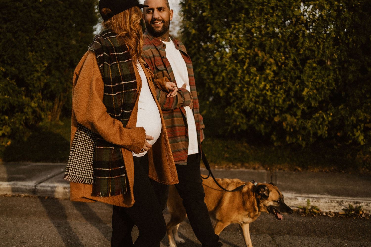 Couple walking a brown dog outdoors in autumn outfits, woman in plaid scarf and man in flannel shirt.