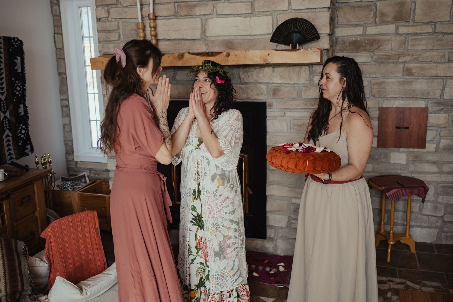 Bride reacts with joy as bridesmaid presents floral crown in rustic stone fireplace living room during wedding preparations.