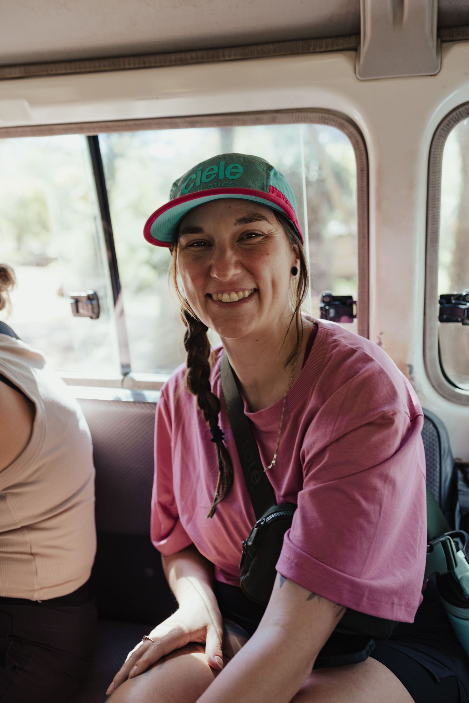 Smiling young woman in pink shirt and colorful snapback cap sitting inside a vehicle with braided hair.