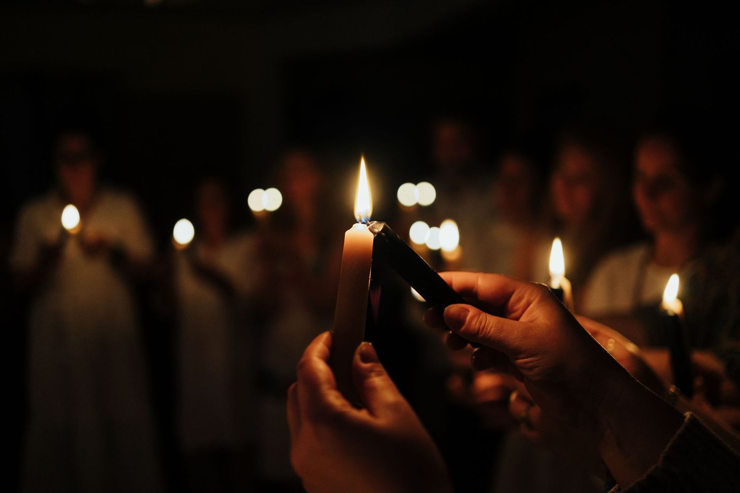 Hands holding lit candles in a dark candlelight vigil ceremony with blurred participants in background.