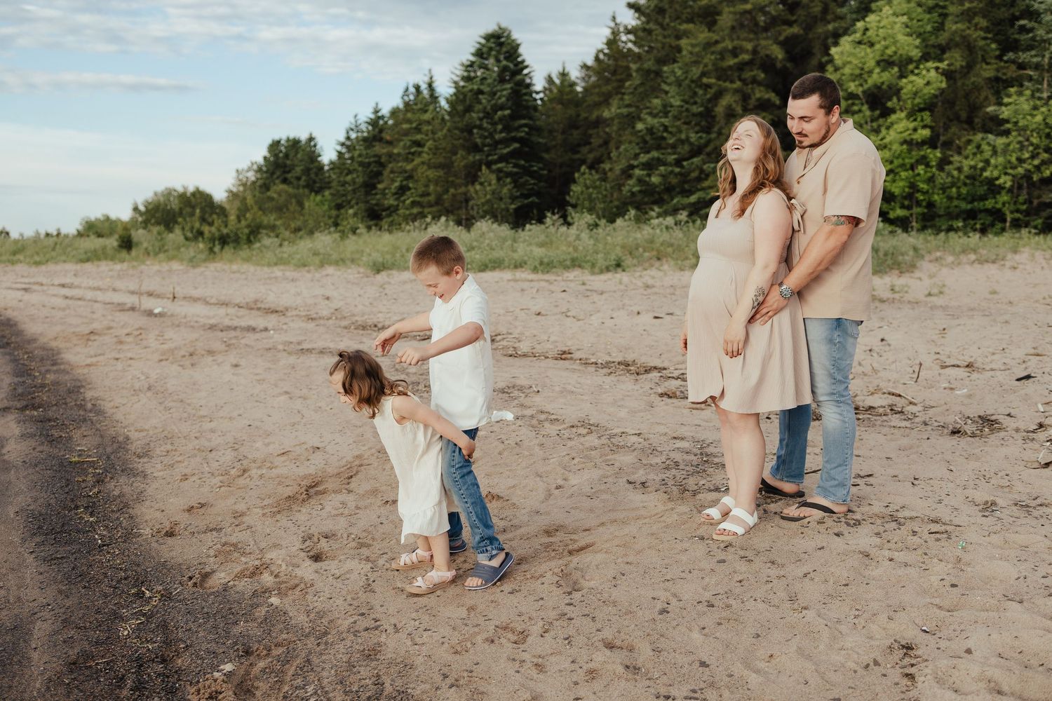 Pregnant woman and man stand on sandy beach while two young children play together in front of tall green trees.