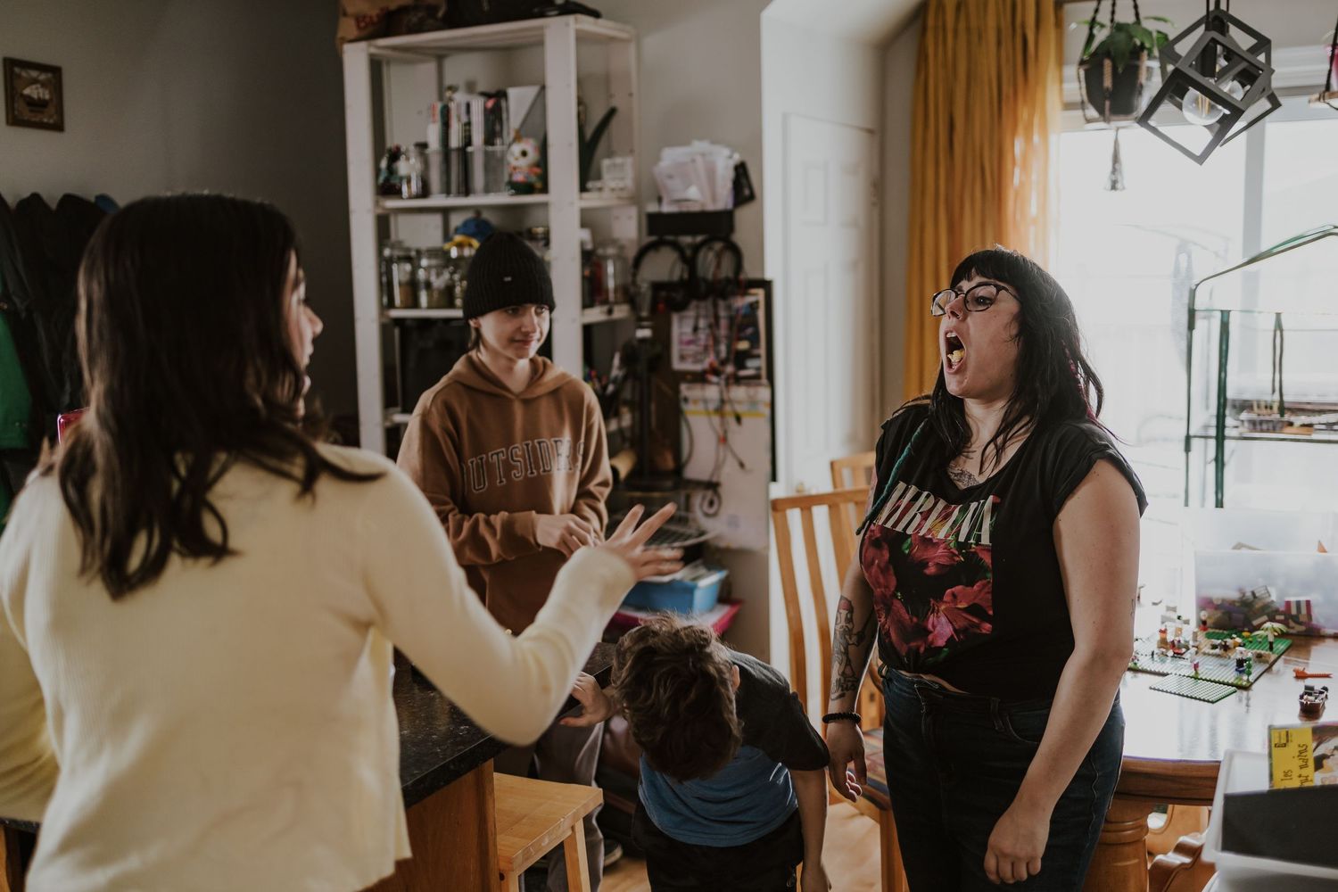 Women laughing and conversing animatedly in a cozy home setting with shelves and plants in the background.