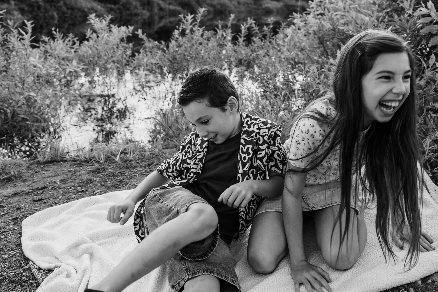 Two children laughing and sitting on a blanket outdoors in a black and white photo.
