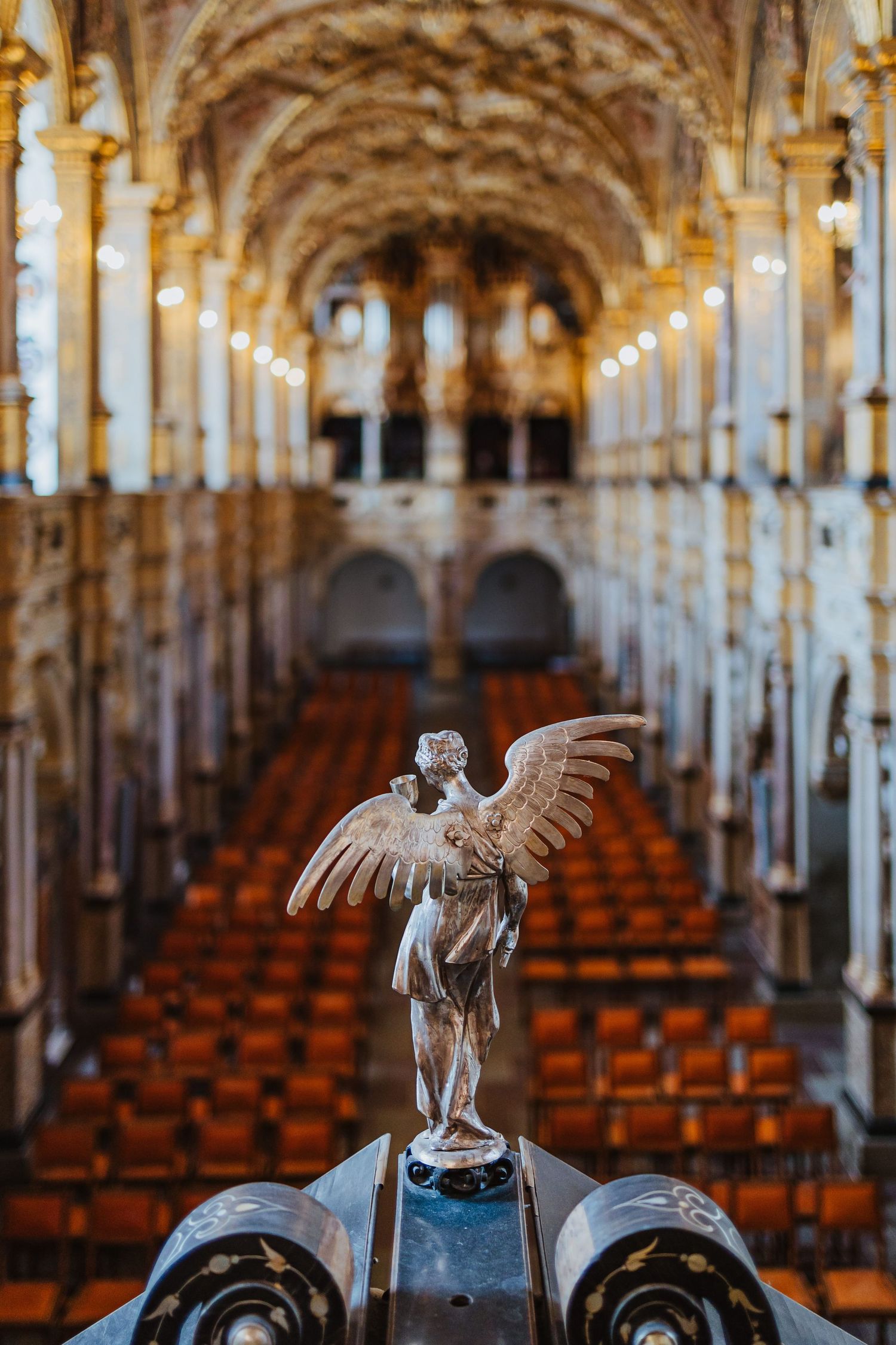 Angel statue with wings in ornate cathedral interior with rows of wooden seats and golden ceiling.