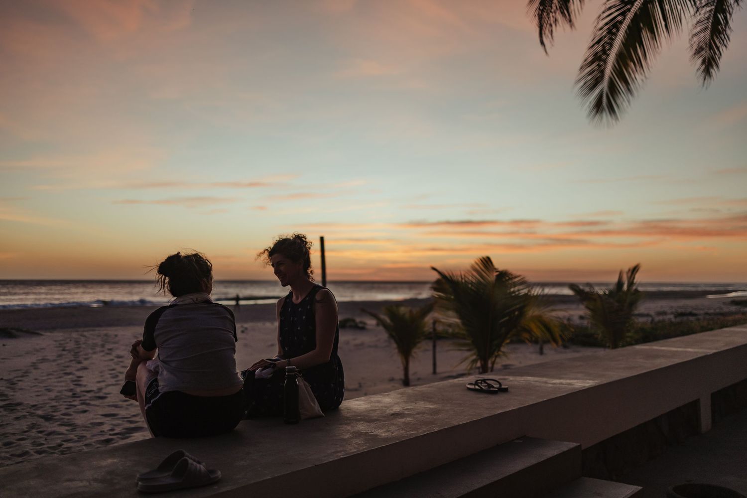 Two people sit on a wall watching a stunning beach sunset with palm trees and colorful sky in the background.