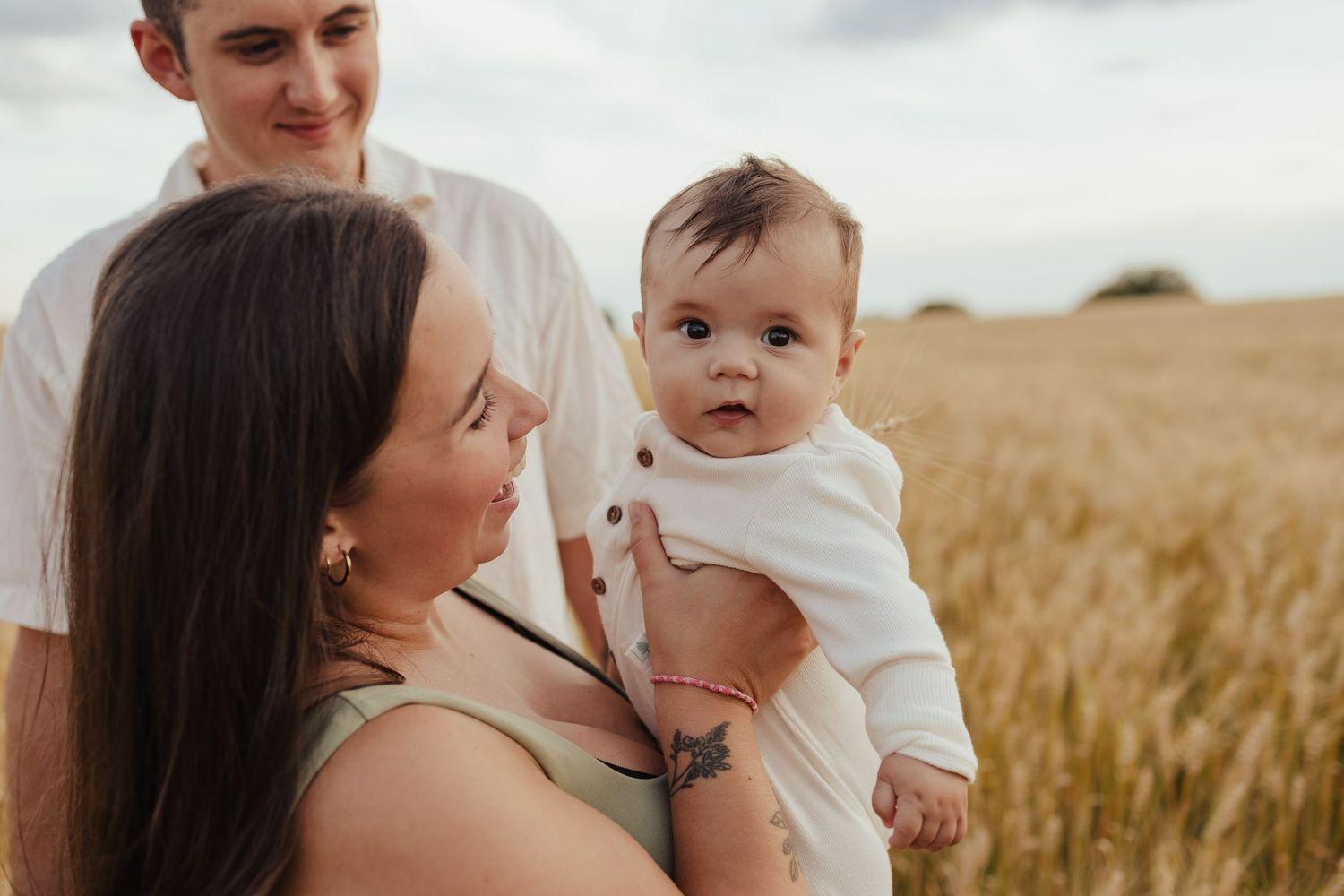 A young family with a baby in a wheat field, mother holding infant in white outfit while father smiles behind them.