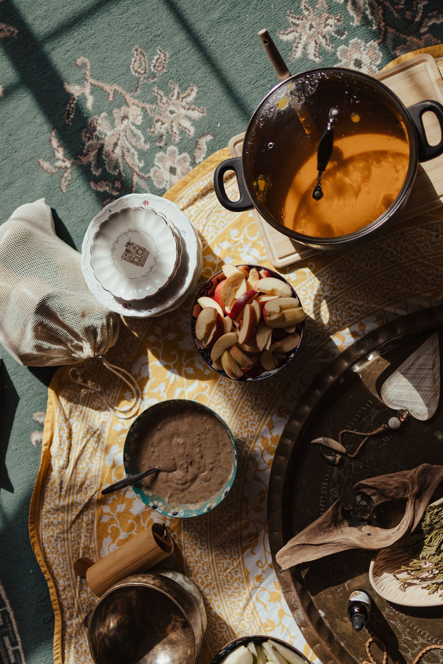 Overhead view of a cozy meal spread with pumpkin soup, chocolate porridge, sliced apples, and tea on a patterned cloth.