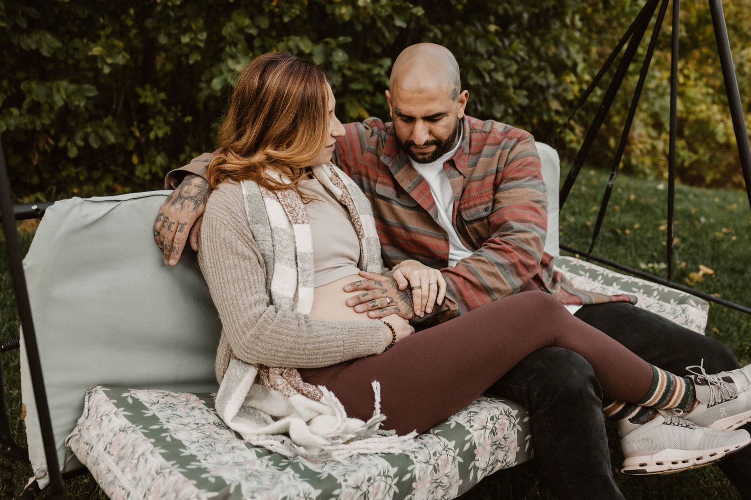 Expecting couple sitting on outdoor swing, man looking at pregnant woman's belly surrounded by nature.
