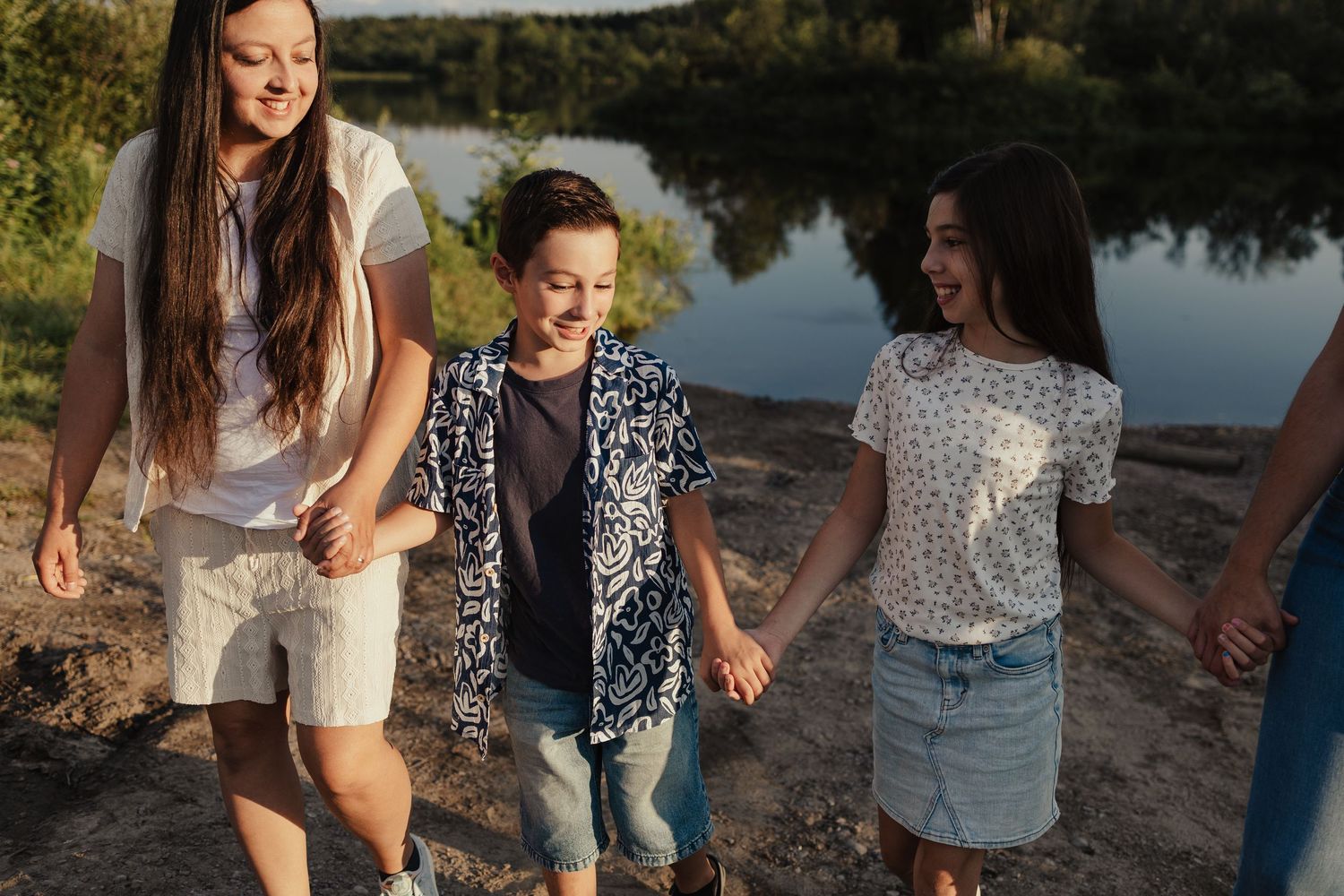 Three happy kids holding hands and walking together near a lake on a sunny day.