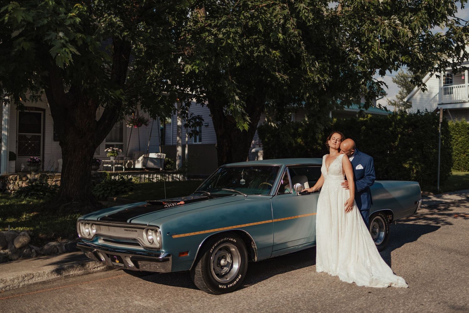 Bride and groom kissing beside a classic vintage blue muscle car on a sunny street on their wedding day.