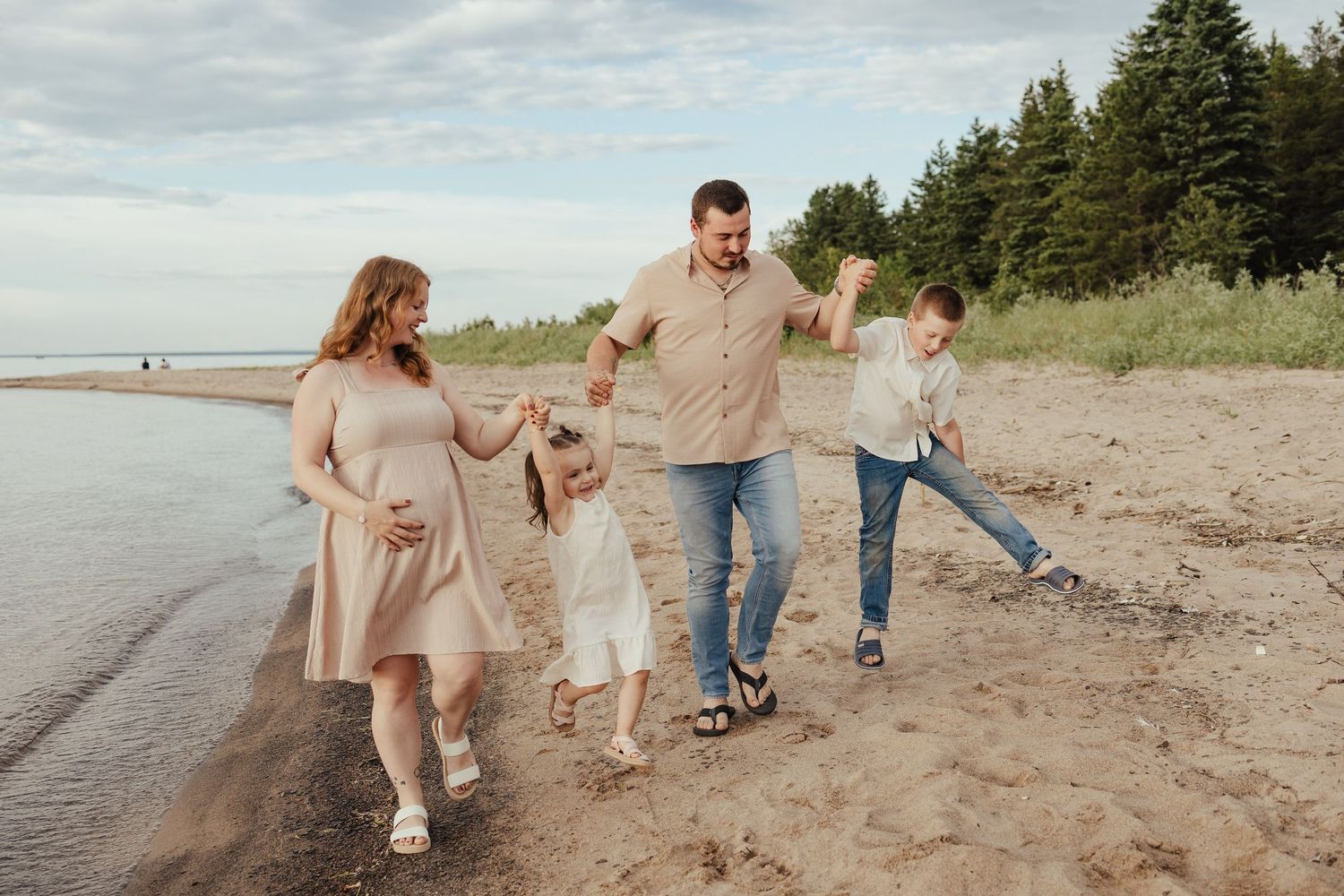 Pregnant woman walks on beach with husband and two children holding hands near shoreline with trees.