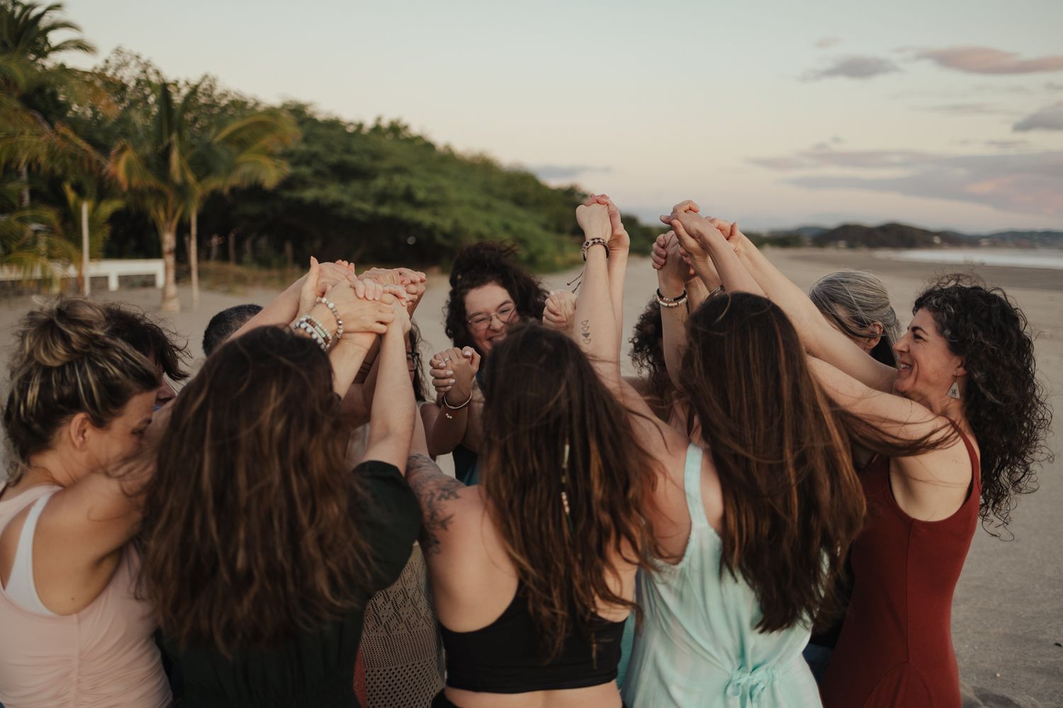 Women holding hands in a circle on a tropical beach at sunset, celebrating together in activewear.