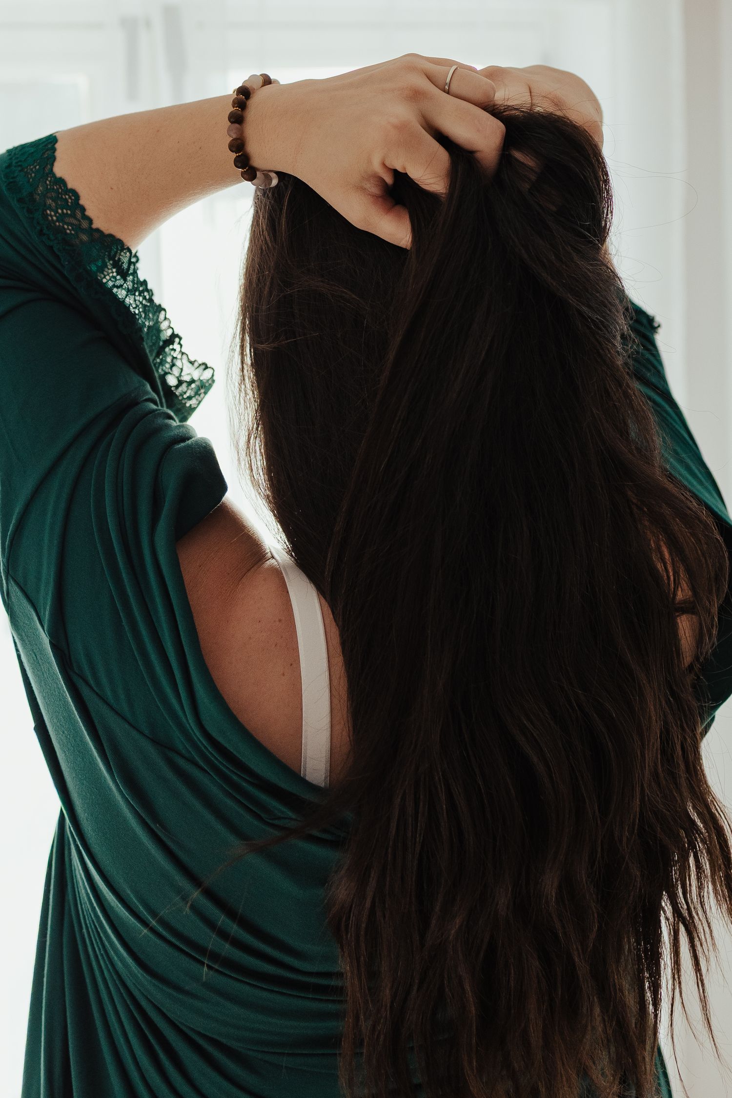Woman in green lace robe with long dark hair, hands raised styling hair, wearing white bra and beaded bracelet.