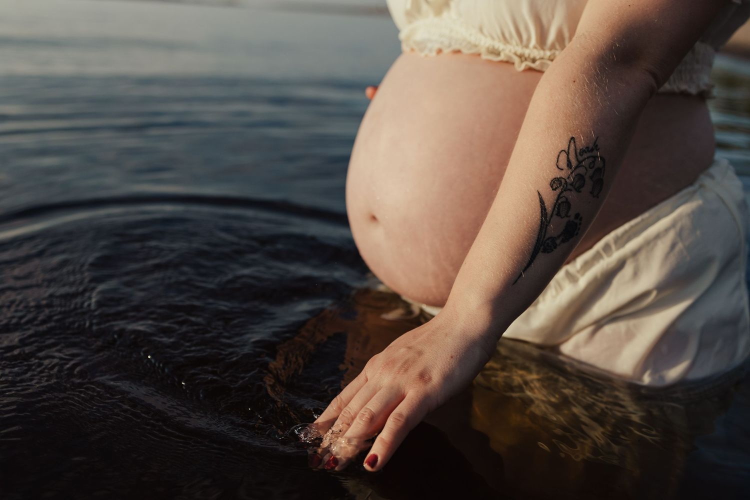 Pregnant woman with tattoo standing in water, wearing white dress, showing baby bump near rocky shore.