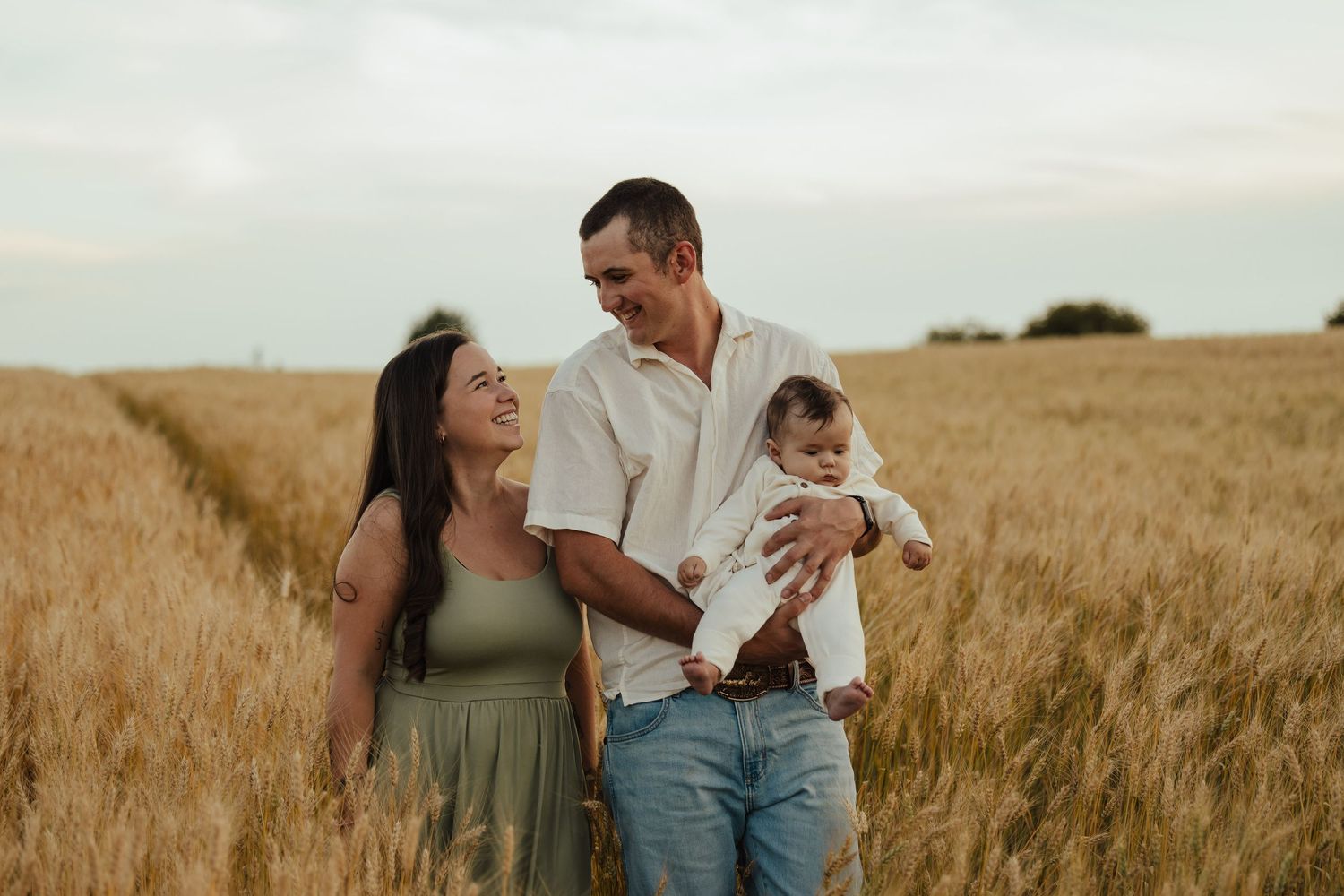 Happy family with baby posing in golden wheat field, parents smiling outdoors in warm countryside setting.