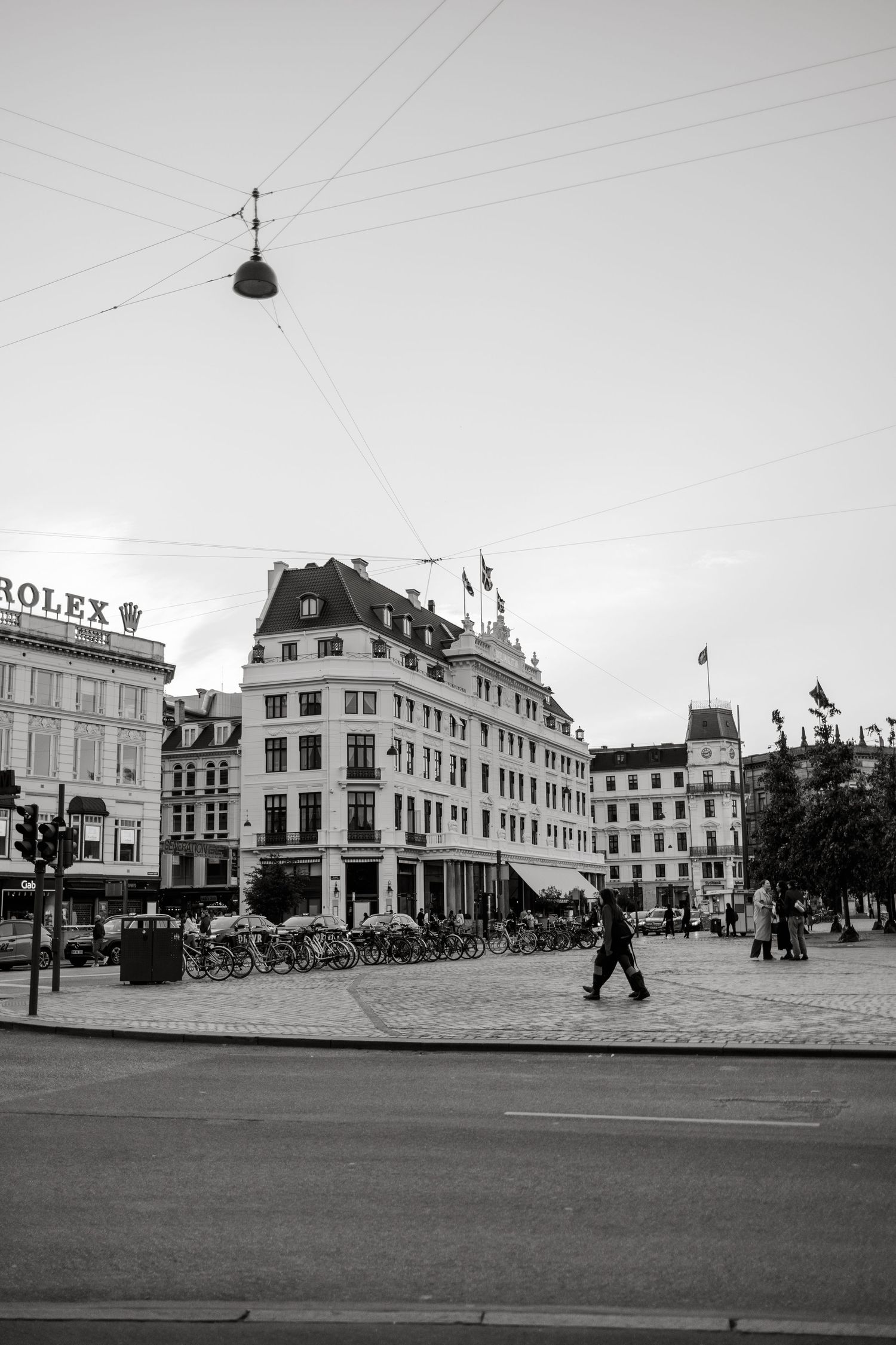 Black and white photo of a European city square with historic buildings, cyclists, pedestrians, and a Rolex sign visible.