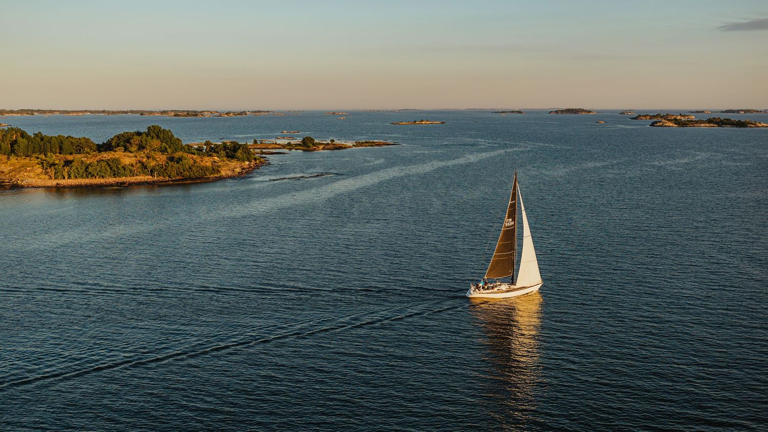 A white sailboat glides through calm blue waters surrounded by small rocky islands at golden hour.