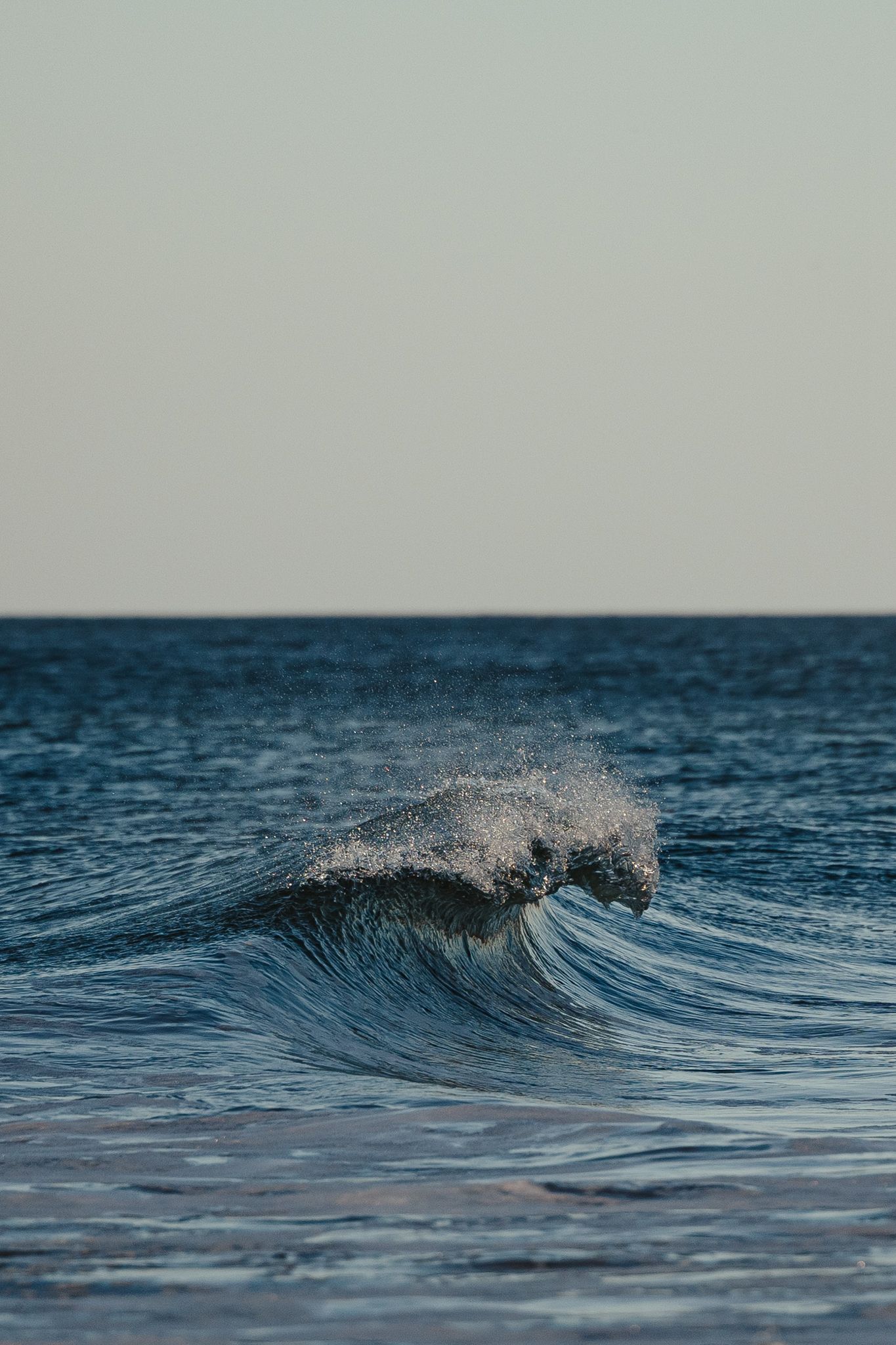 A powerful ocean wave crests and splashes against calm blue sea water under a clear sky.
