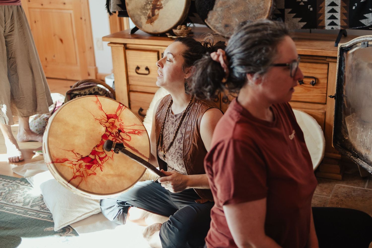 Two women playing large frame drums in a cozy room during a shamanic drumming circle ceremony.