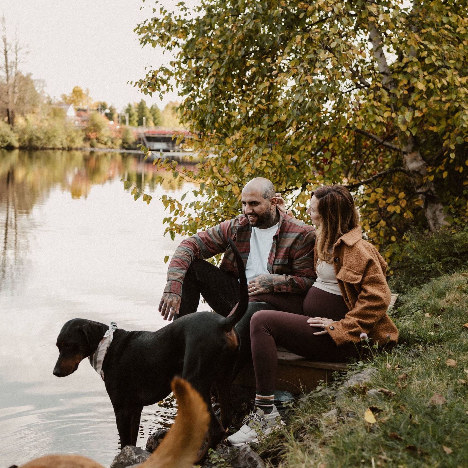 Couple sitting by a tranquil river with their black dog on an autumn day, surrounded by golden foliage.