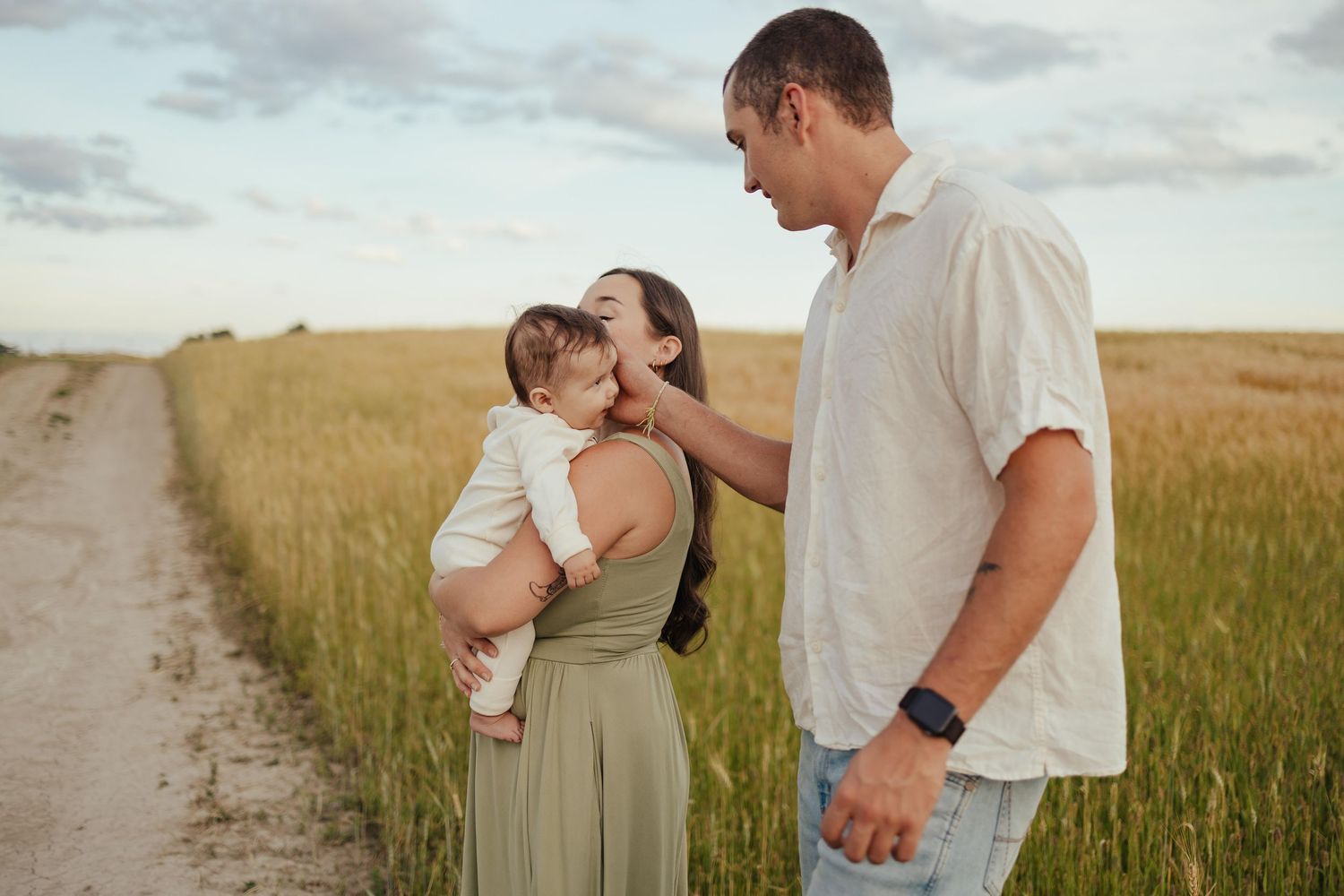 A young family with a baby stands on a dirt path in a green field at sunset, sharing a tender moment.