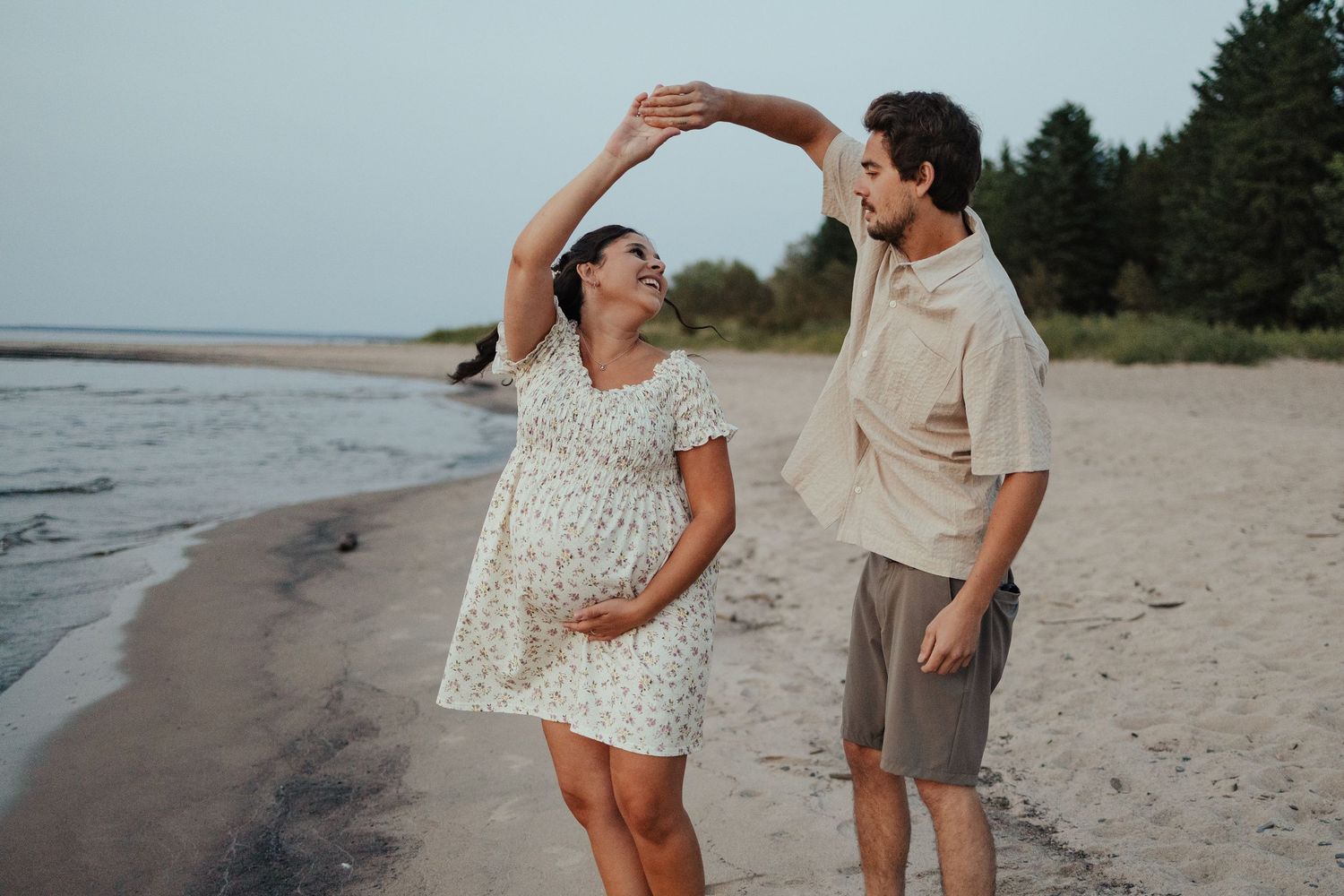 Pregnant couple dancing on beach shore, man spinning woman in floral dress, trees and water in background.