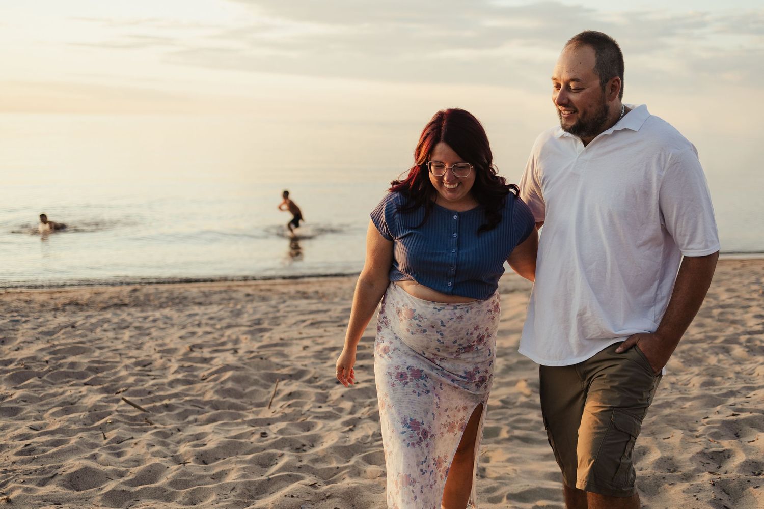 Couple walking on sandy beach at sunset, smiling and holding each other, with ocean waves and a surfer in the background.