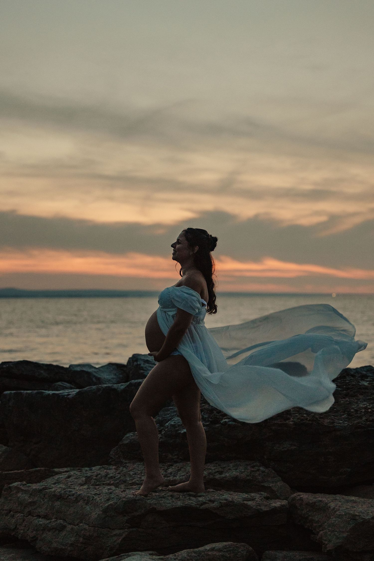 Pregnant woman in flowing white dress standing on rocks at sunset by the ocean.