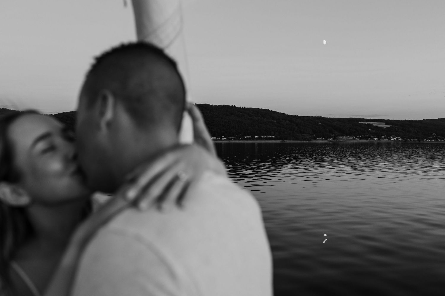 Black and white photo of a couple sharing a romantic kiss by a serene lake at dusk with hills in the background.