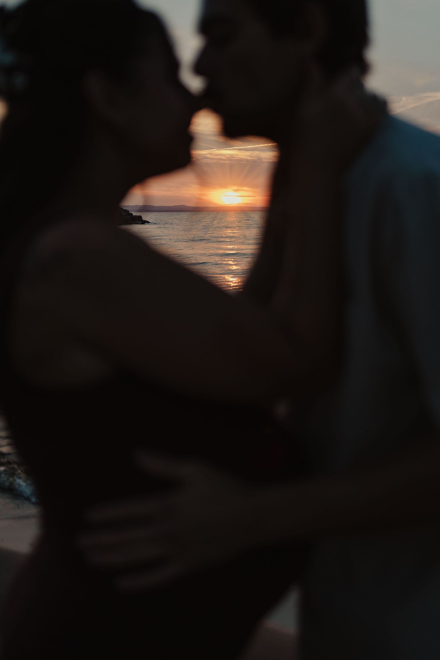 Romantic couple silhouetted against a vibrant ocean sunset, sharing a kiss on the beach at dusk.