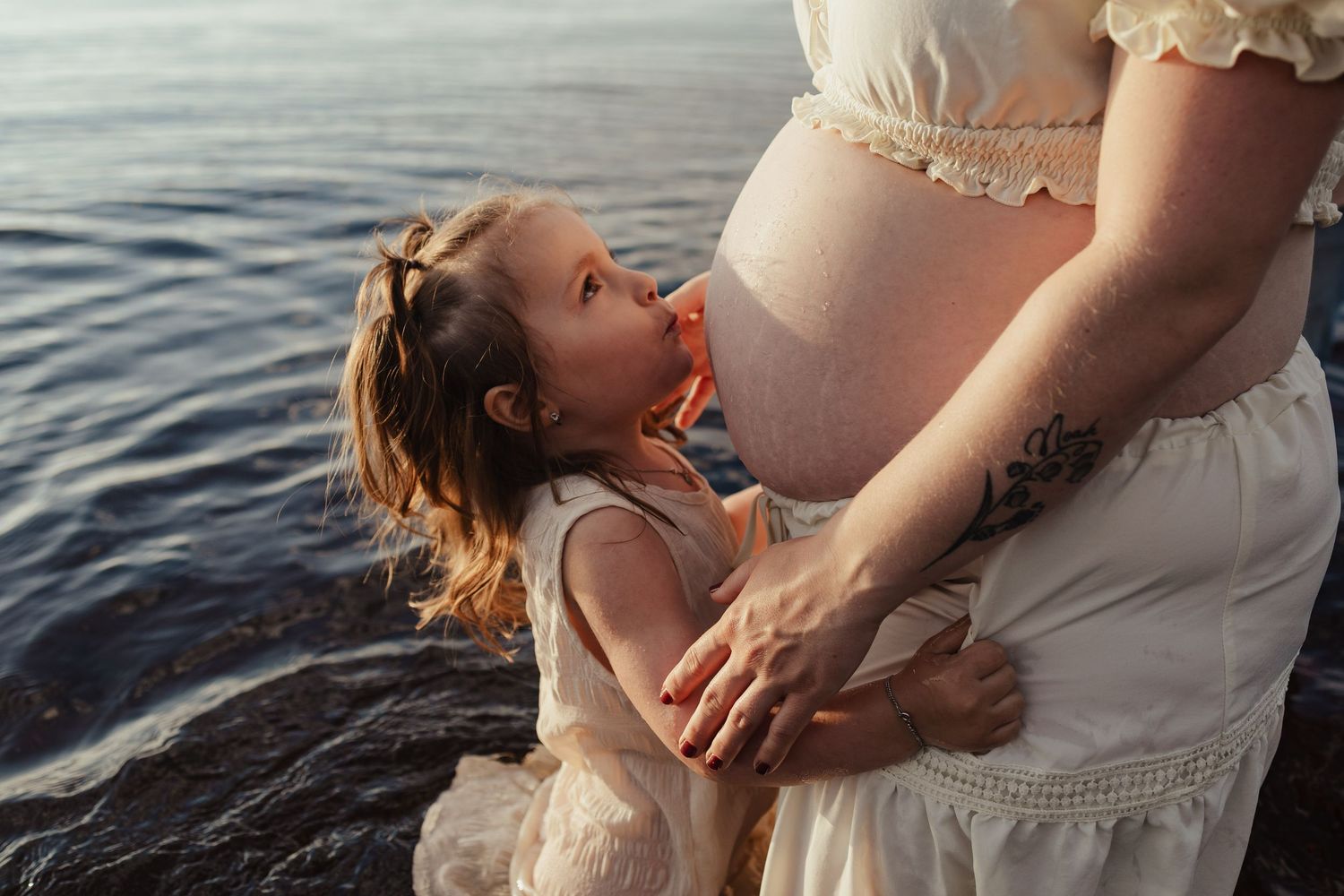 Young girl kissing pregnant mother's belly while standing in shallow water.