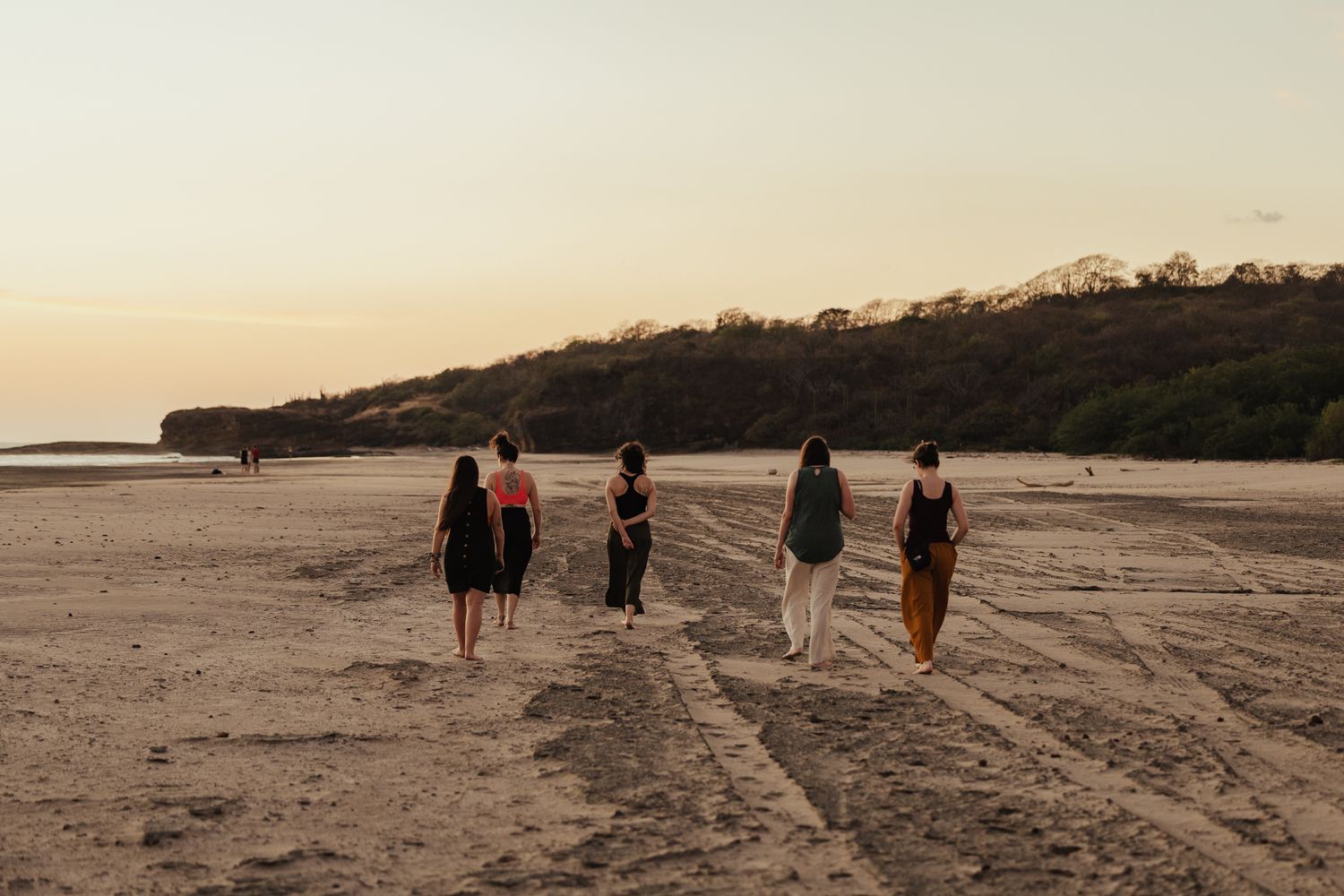 Four women walking along a sandy beach at sunset with lush green hills in the background.
