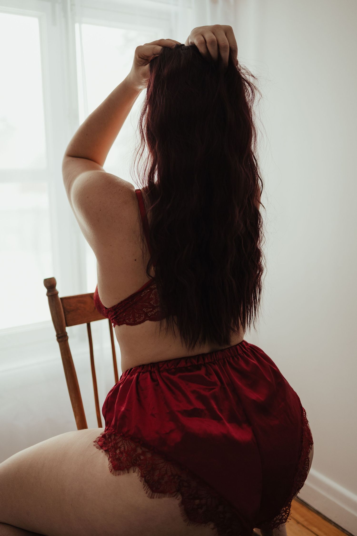 Woman with long dark hair in red lace lingerie sitting on a chair facing a bright window.
