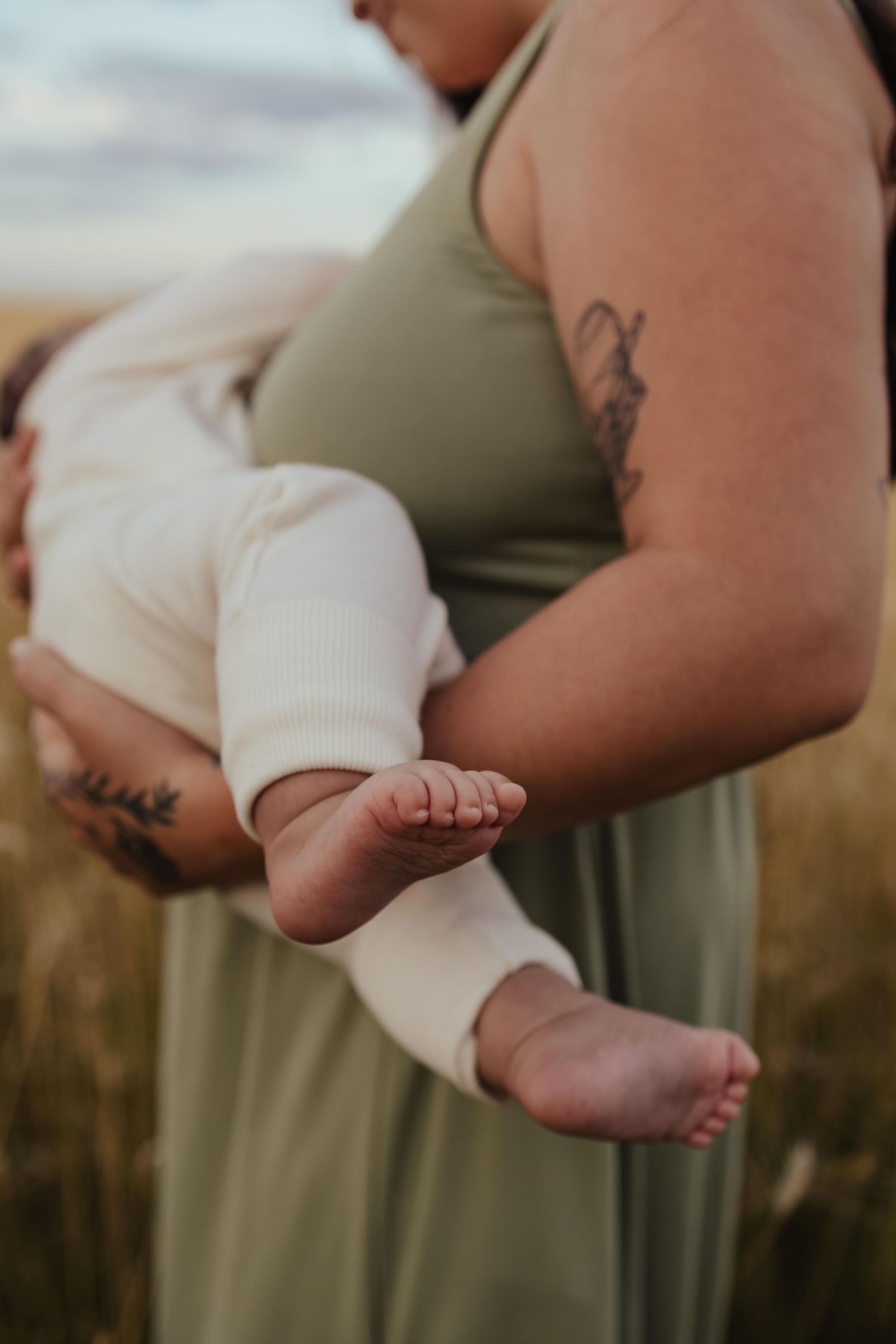 Mother in olive dress with tattoos cradling newborn baby in white onesie outdoors in golden field.