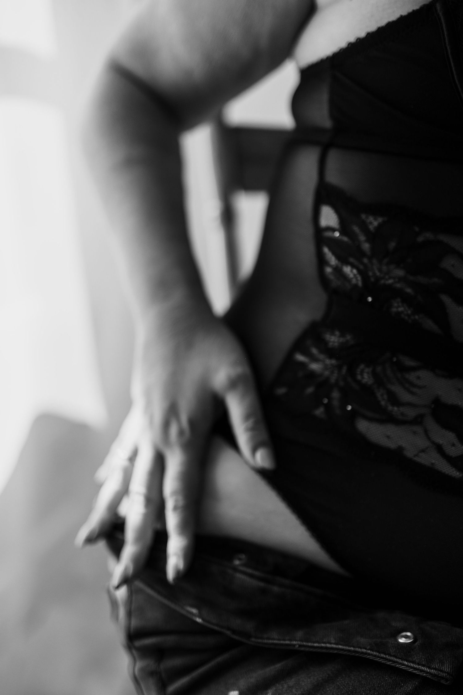 Black and white photo of a woman in lace lingerie and jeans, hand resting on hip near waistband.