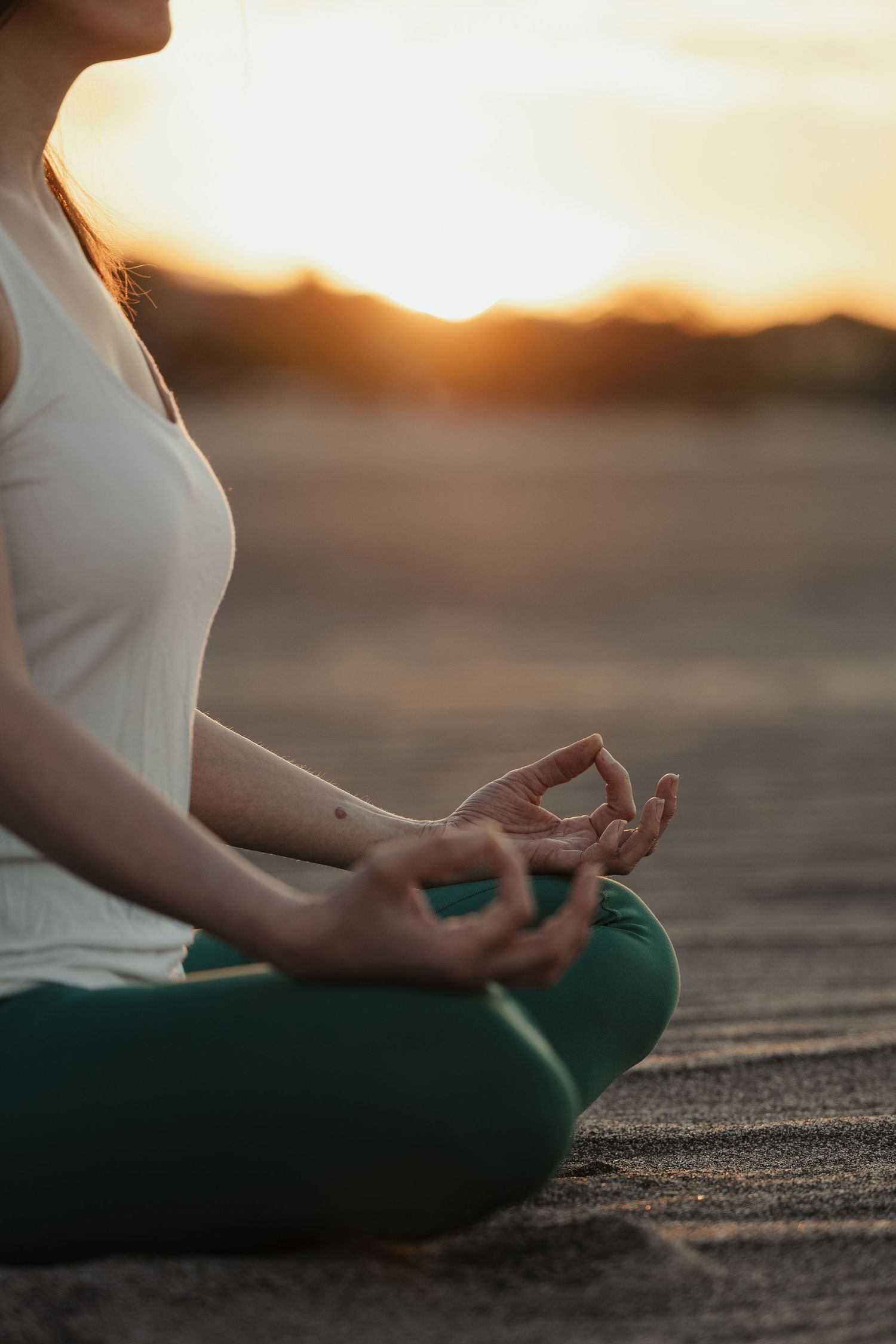 Woman meditating in lotus pose at sunset, hands in mudra gesture, wearing white top and green yoga pants outdoors.