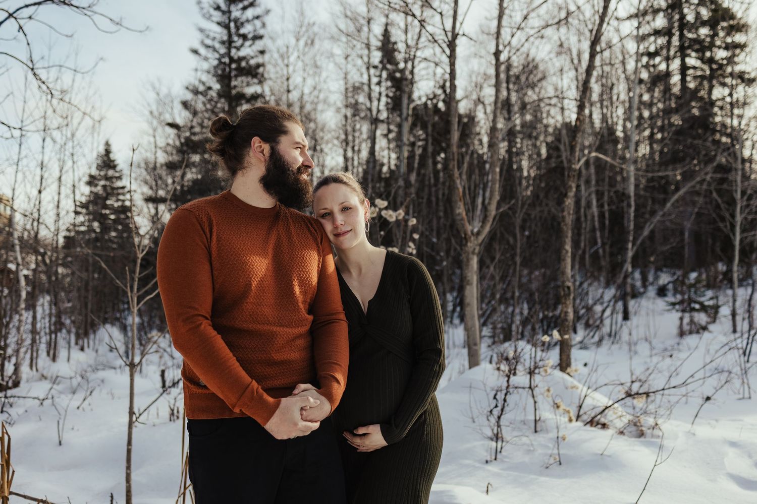 Couple embracing in snowy winter forest during maternity photo session, surrounded by bare birch trees.