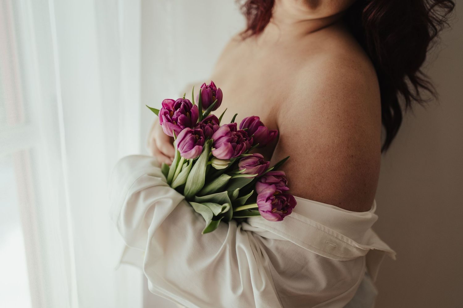 Woman in white off-shoulder wrap holding a bouquet of purple tulips near a bright window.