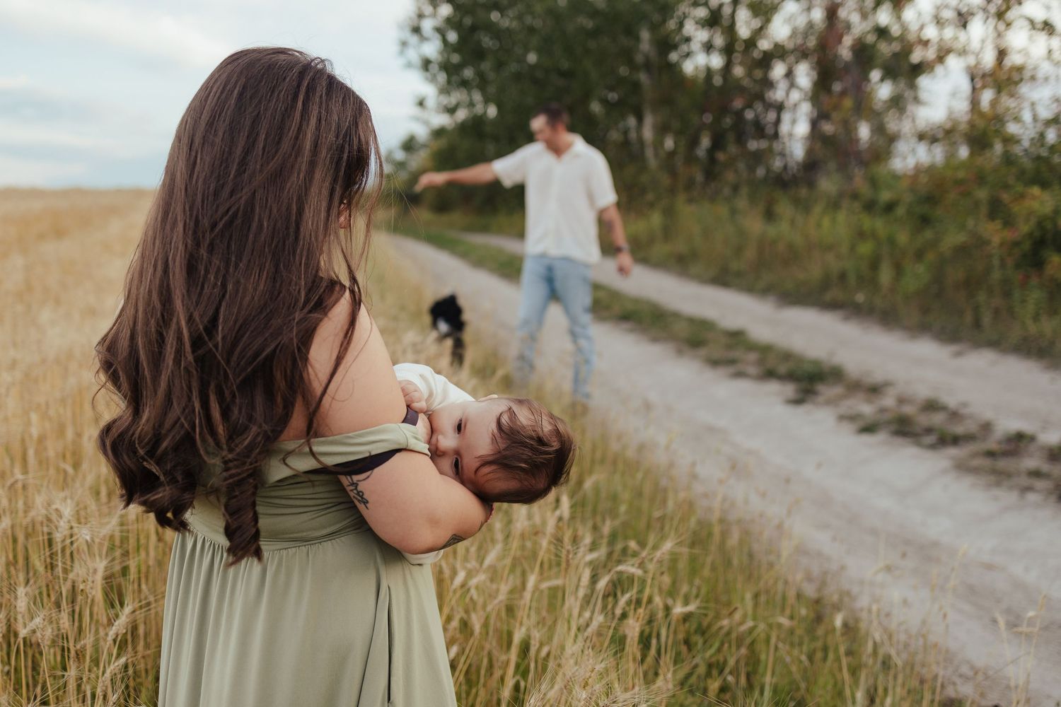Mother in green dress cradling newborn baby in a field while father and dog walk along a dirt path.