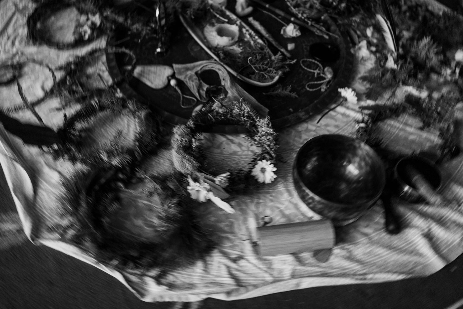 Black and white overhead shot of ceremonial items including bowls, flowers, and natural objects arranged on a cloth.