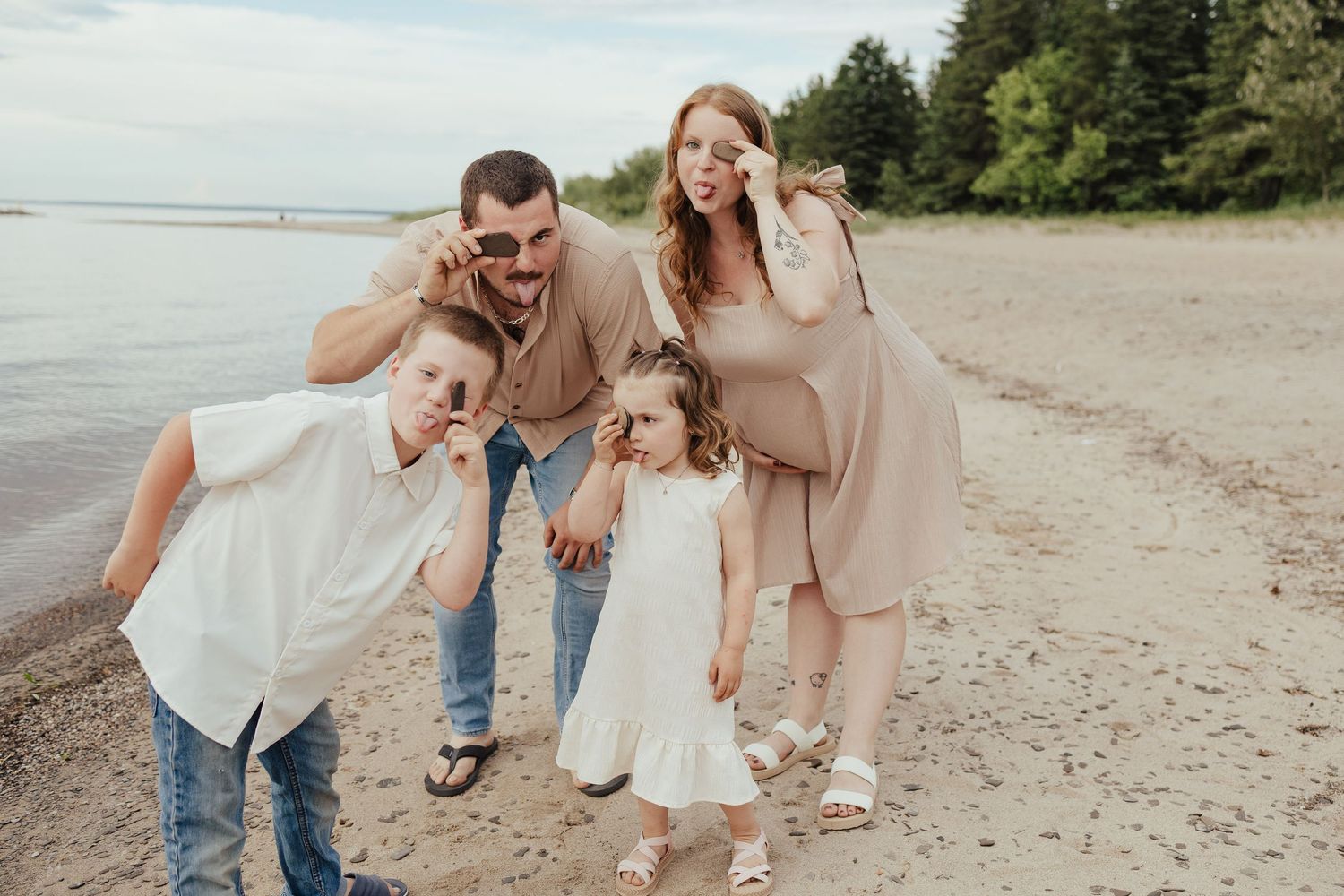Pregnant woman and family posing playfully on a sandy beach with trees in the background.