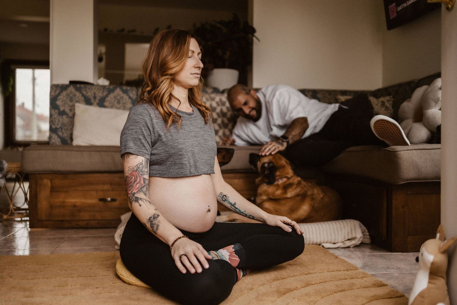 Tattooed pregnant woman meditating on floor while partner plays with dog on couch behind her.