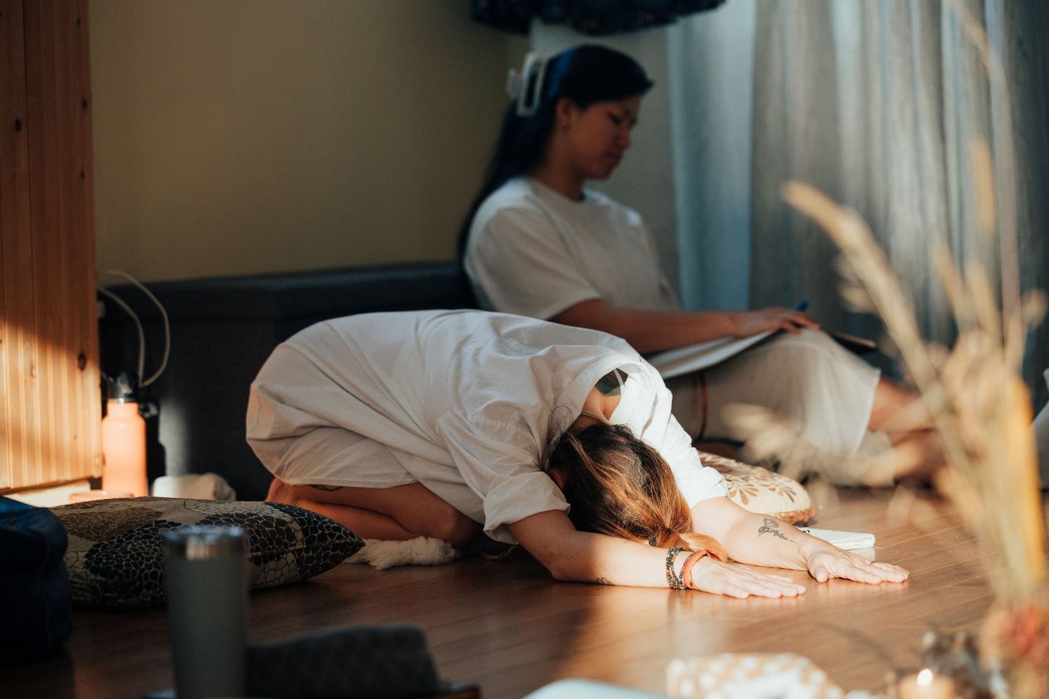 Two women in white robes relaxing in a cozy spa room with warm sunlight streaming through the window.